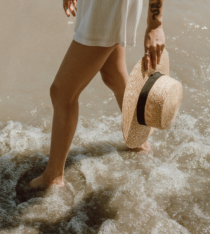 a woman walking on the beach carrying a straw hat