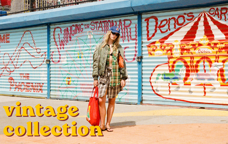 A woman stands on a sidewalk in front of closed blue roller shutters with colorful graffiti, including a carousel mural. She is wearing a green jacket, plaid dress, and carrying a red tote bag.