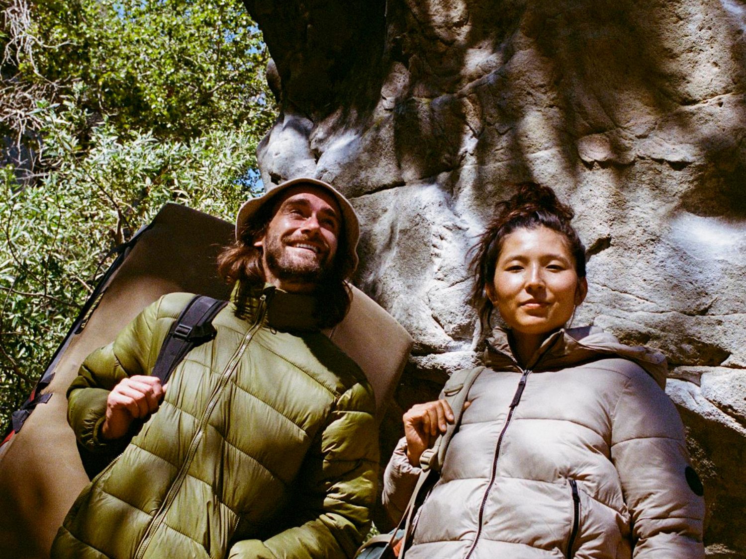 Two people in outdoor clothing standing in front of a rocky wall with trees in the background