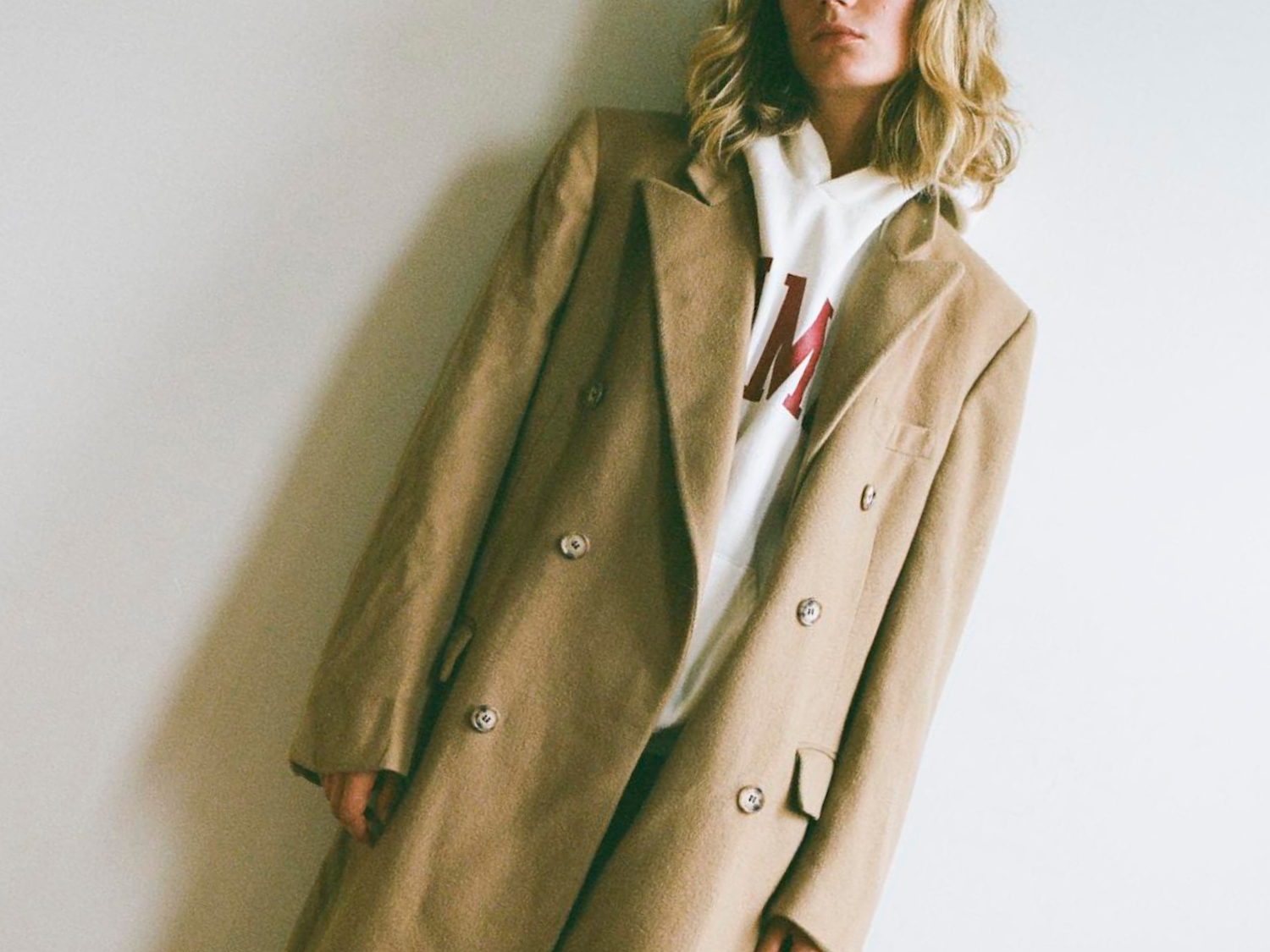 Person wearing a long beige coat over a white shirt with red lettering against a plain background
