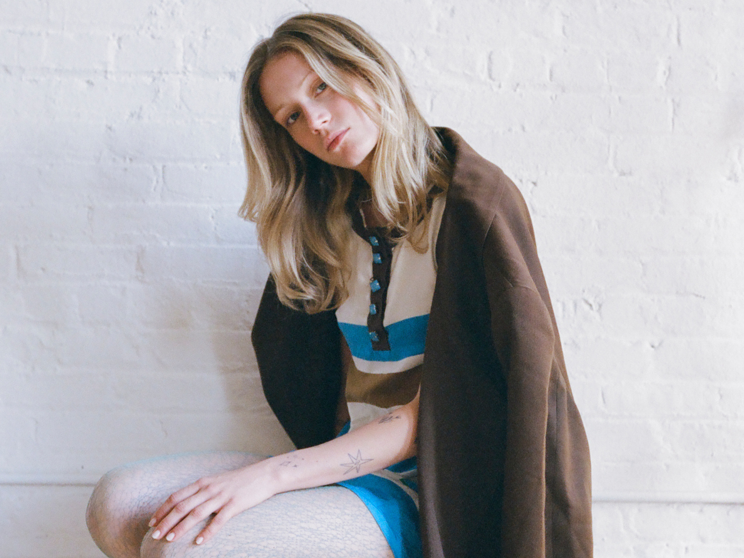 Woman sitting on a couch wearing a brown coat against a white brick wall.