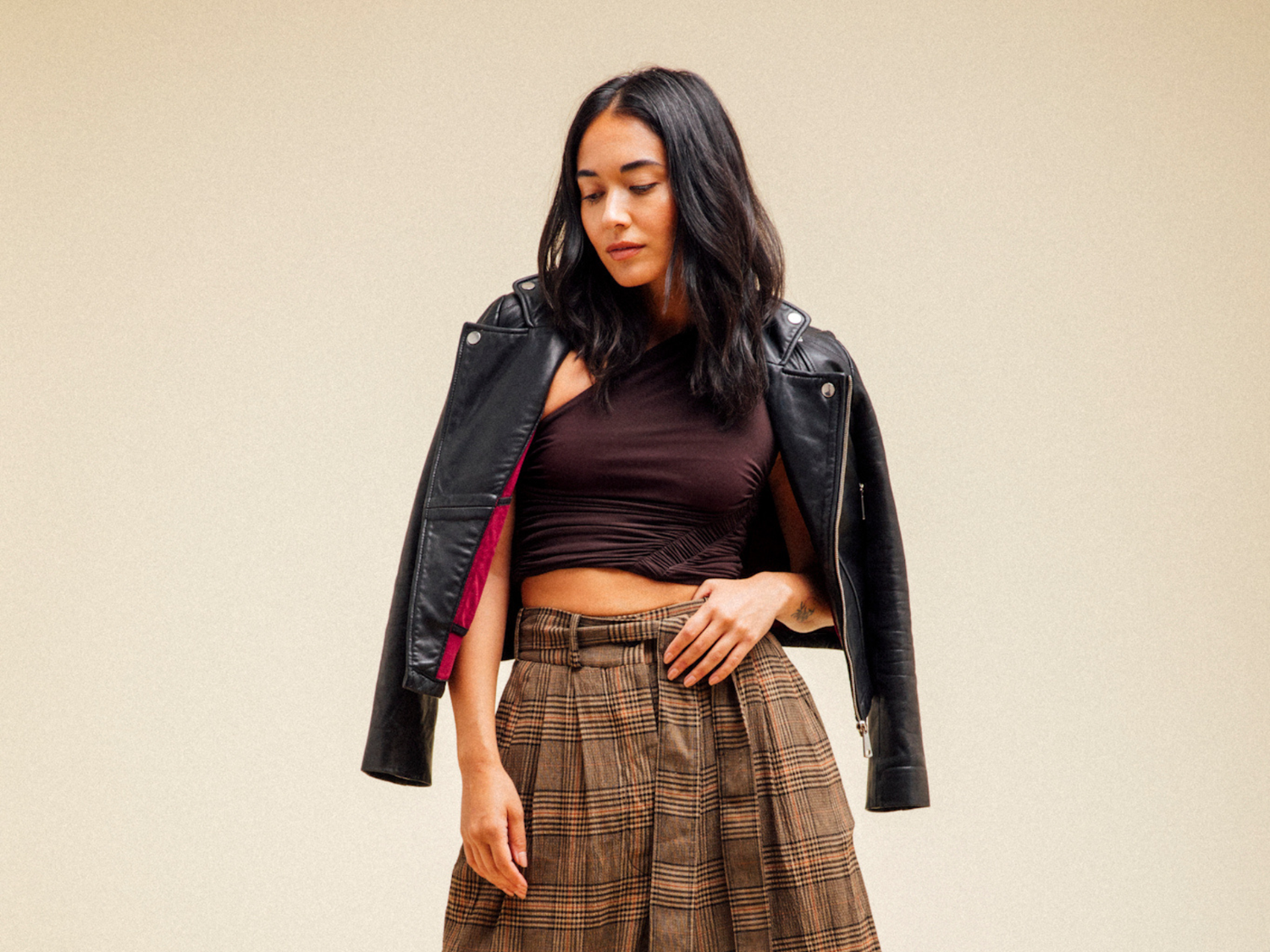 Woman wearing a black leather jacket, brown top, and plaid skirt against a beige background