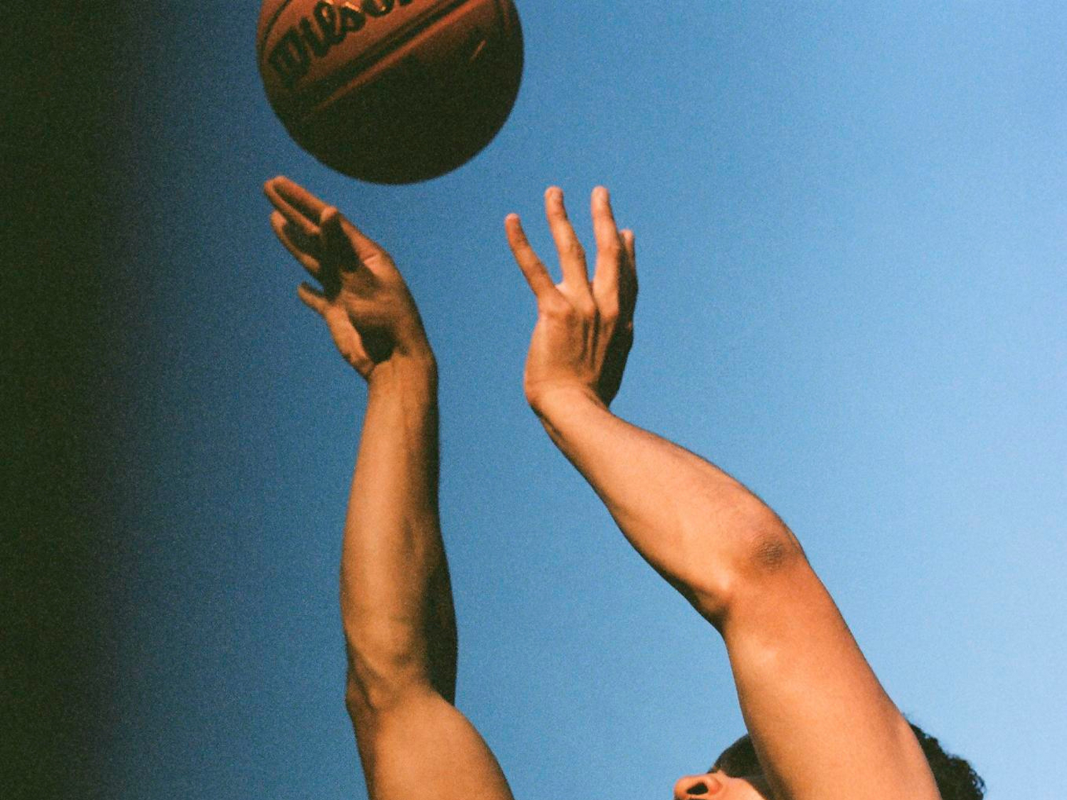 Two hands reaching up towards a basketball against a blue sky.