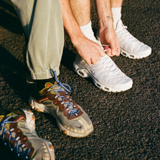 A close-up lifestyle shot of a person sitting on asphalt, tying the laces of a white Nike sneaker. Another pair of multicolor sneakers is visible in the foreground.