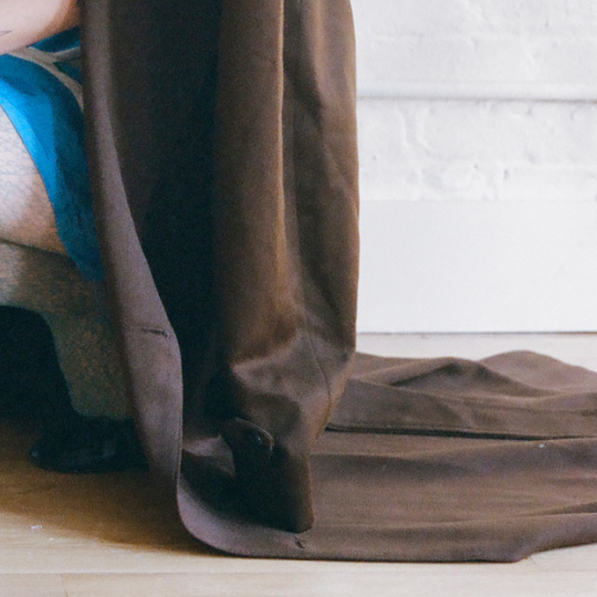 A close-up of a person wearing a long brown coat or dress, with the fabric draped over their lap and pooling on the floor against a white wall.