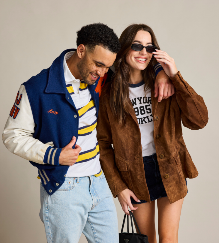 A man and a woman posing together in a studio setting. The man wears a navy and cream varsity jacket over a yellow and white striped polo shirt, paired with light blue jeans.