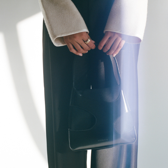 A person in a cream-colored coat and black trousers holds a black leather Hermès handbag with a transparent black pouch insert, set against a bright, sunlit white background.