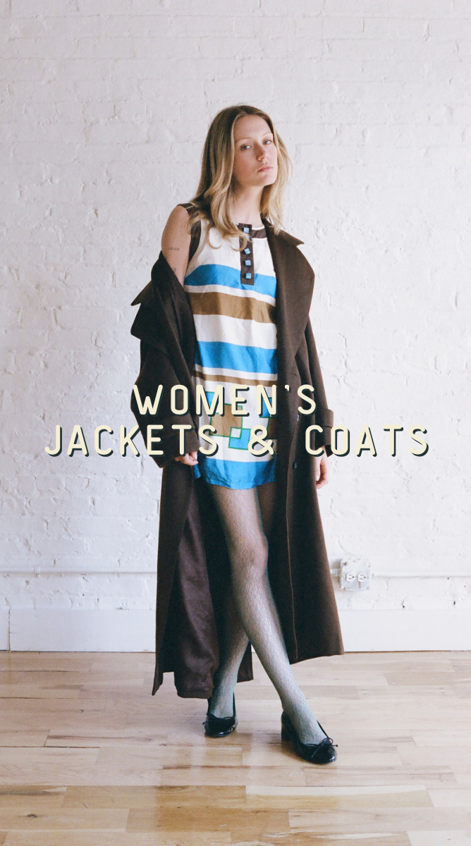 a woman wearing a dress with long brown coats standing in front of a brick wall. Caption: "Women's Jackets & Coats"